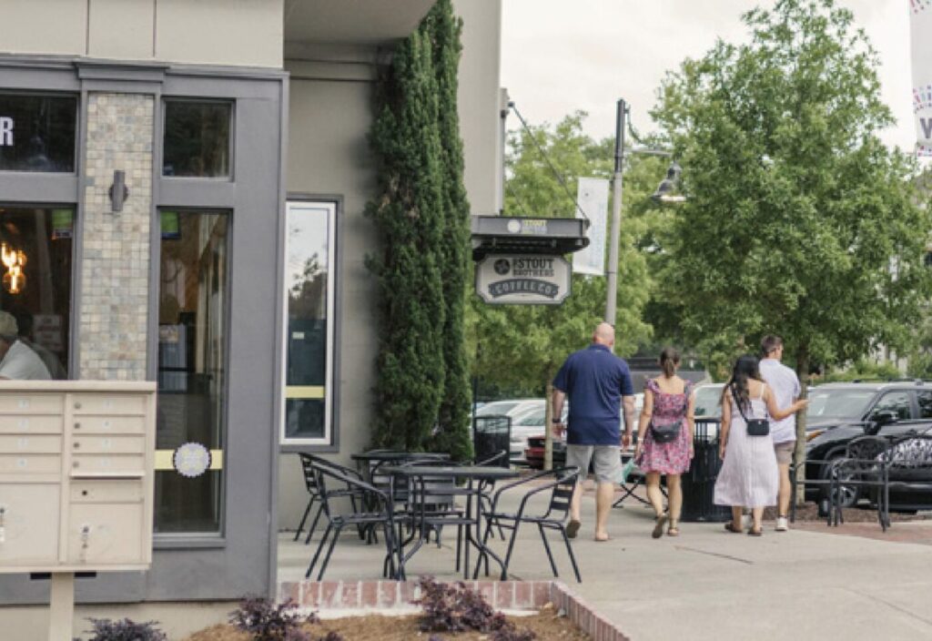 People walk past an outdoor patio and storefronts along a sidewalk lined with parked cars.
