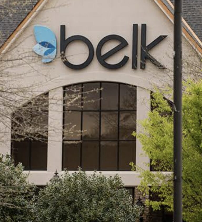 Belk store sign on the exterior of a shopping center building partially framed by trees.
