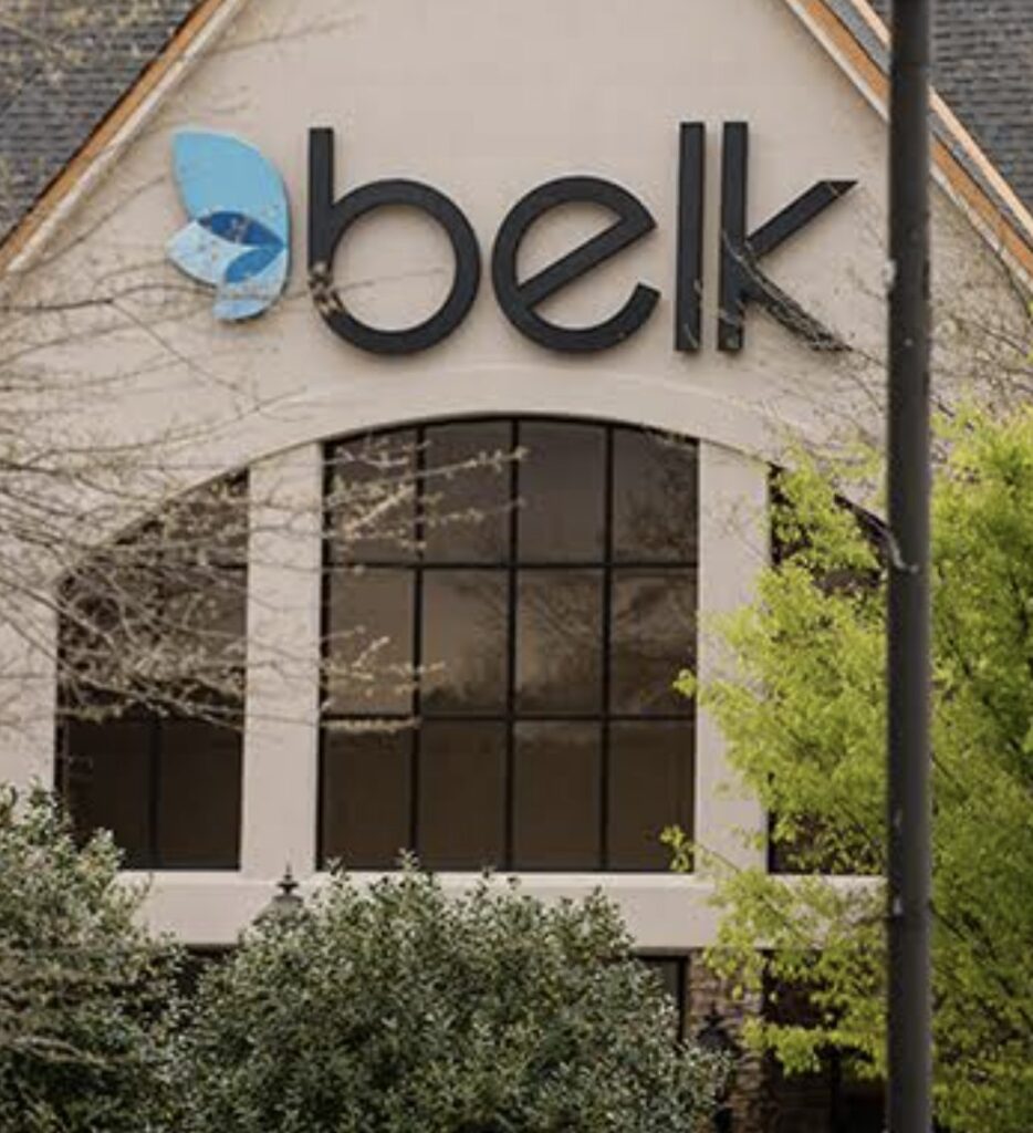 Belk store sign on the exterior of a shopping center building partially framed by trees.