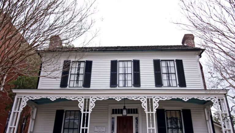 White two-story historic house with a front porch, black shutters, and bare tree branches in front