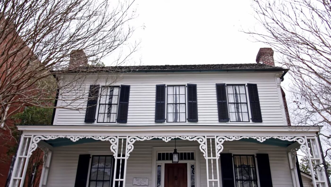 White two-story historic house with a front porch, black shutters, and bare tree branches in front
