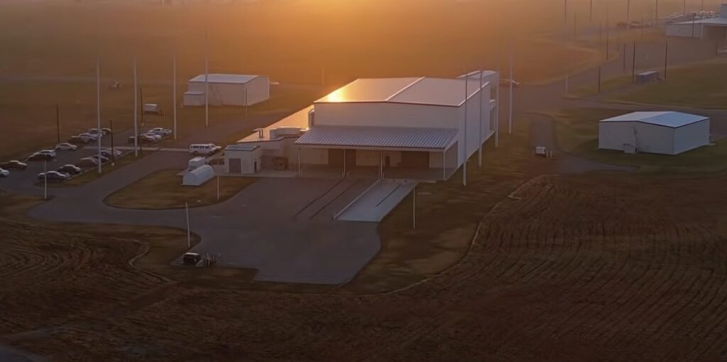 A small industrial building and paved lot at sunset, with utility poles and open fields around it.
