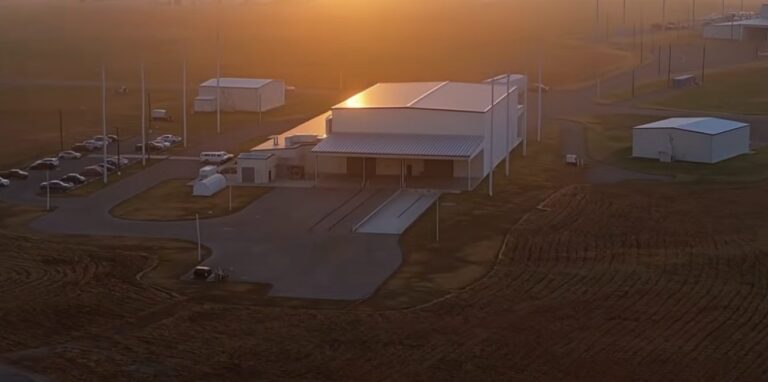 A small industrial building and paved lot at sunset, with utility poles and open fields around it.
